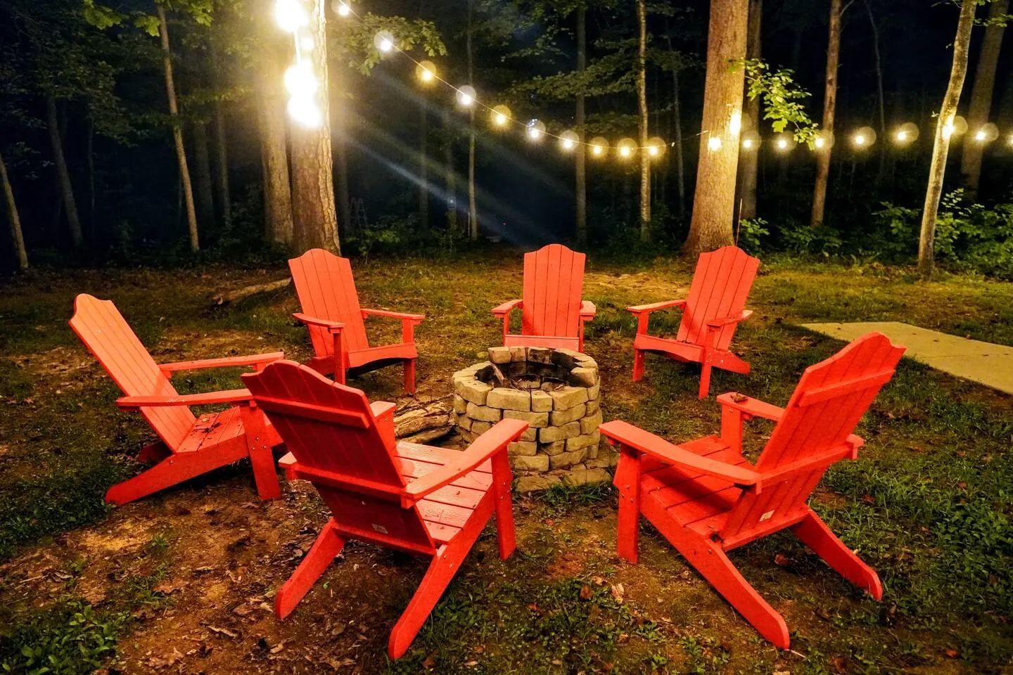 Stone fire pit ringed by red Adirondack chairs under 200 feet of string lights at night